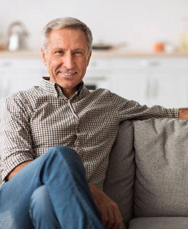 Älterer Mann mit grauen Haaren sitzt entspannt auf einem Sofa und lächelt in die Kamera.