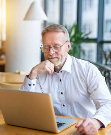 Älterer Mann mit Brille und Bart sitzt nachdenklich vor einem Laptop in einem modernen, hellen Büro mit großen Fenstern.