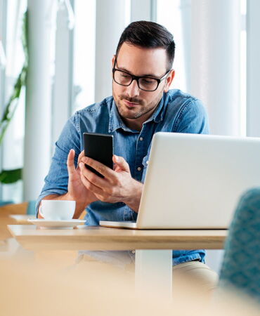 SSZ Beratung Definition Mobile Work Mann mit Brille und Jeanshemd sitzt in einem Café mit Laptop und Kaffeetasse, konzentriert auf sein Smartphone blickend.
