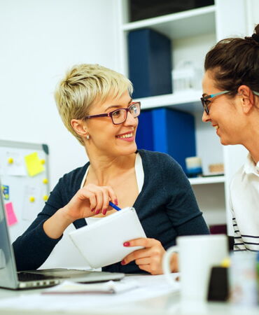 Zwei Frauen mit Brillen unterhalten sich lachend in einem modernen Büro, eine hält Notizbuch und Stift.