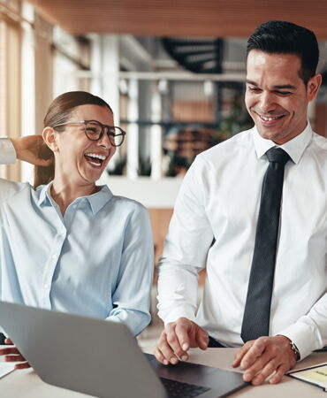 Lachende Frau und Mann im Business-Outfit arbeiten gemeinsam am Laptop.